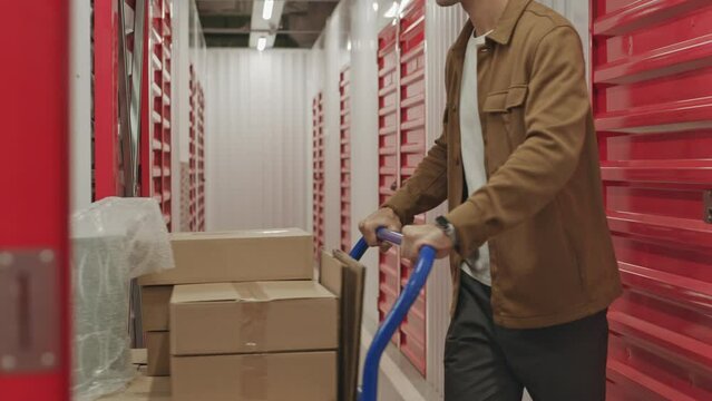 Man walking with dolly cart loaded with boxes through corridor in self service warehouse while going to his unit