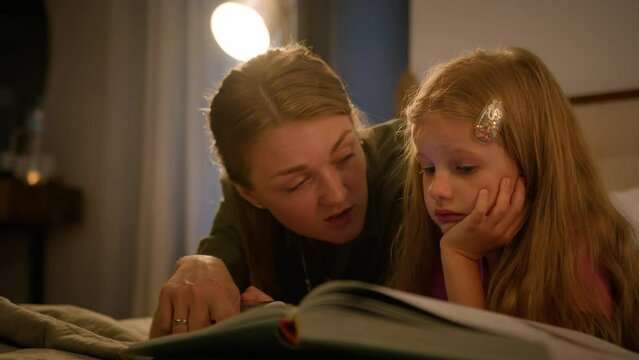 Happy Caucasian family loving caring mother reading book to little kid daughter using flashlight in bedroom lying on bed evening mom and child girl enjoying read fairytale story bedtime parenthood