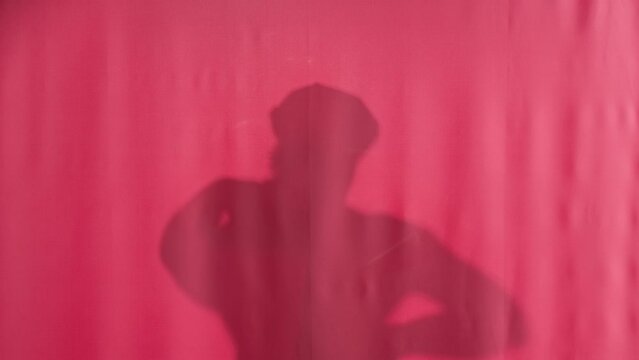 Silhouette of a man washing behind a pink shower curtain. The man pulls back the curtain smilingly looking straight ahead and closes the shower curtain again.