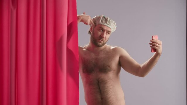 A man takes selfies using a smartphone while showering. Nude man wearing waterproof cap, near pink shower curtain poses in studio on blue background close up.