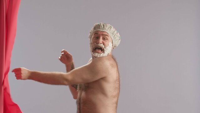 A man with shaving foam on his beard peeks out from behind a pink shower curtain and screams in fright. Male with bare torso and wearing a shower cap. Humorous Beauty Concept.