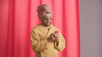 A man is typing a message on his smartphone after a shower, chatting. A man in a robe, with a pink towel on his head and hydrogel patches under his eyes against the background of a pink shower curtain