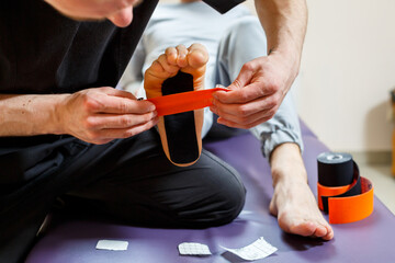 Male physiotherapist doctor massages the feet of a relaxed man sitting on a stretcher. He uses kinesio tape. Taping the foot
