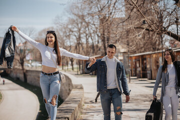 Three beautiful people having fun while hanging out in the city. Lovely brunette girl walking on the wall by the quay, her male friend is holding her hand to make sure she is not falling down - obrazy, fototapety, plakaty