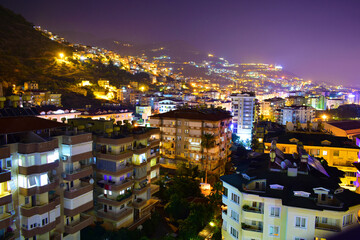 City view of multi-storey modern cottages on a mountainside in the night with colorful lights. Turkey, Alanya, July 2023.