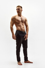 Fototapeta premium Full-length image of handsome young man with muscular relief body standing shirtless in pants isolated over white studio background. Concept of male beauty, body care, fitness, sport, health