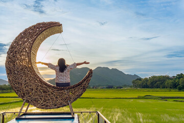 Woman sit on crescent moon chair made of rattan for relaxation on bridge in paddy field with beautiful scenic in evening. Decorative wooden moon furniture as sitting chair for viewpoint in rice field