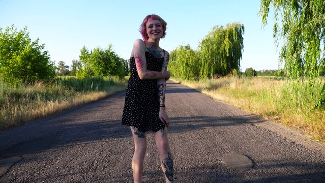 Young hippie woman in black dress walking along empty road at countryside. Happy punk girl with pink hair fooling around while goes on country driveway. Carefree female hipster having fun outdoor