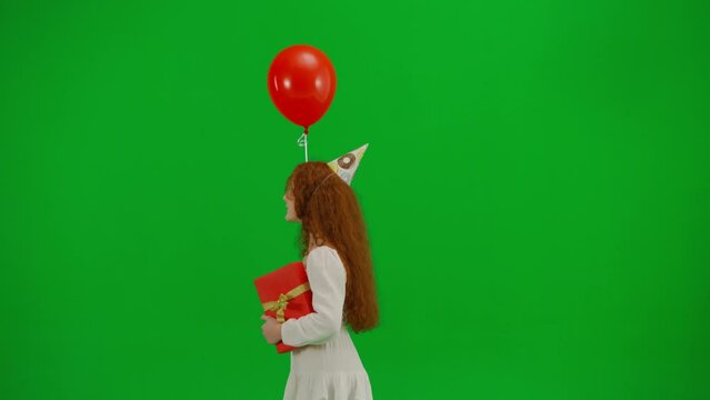 Redheaded little girl in white dress with a festive cap on her head and with a gift in her hands is walking with a red balloon. Green screen. Side view. Concept of holiday, joy and fun.
