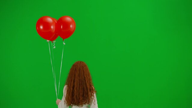 Redheaded little girl in white dress walking with red balloons on green background of studio. Back view. Concept of holiday, joy and fun.