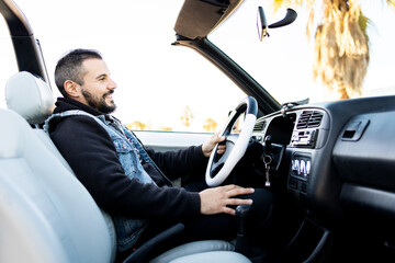 Profile view of an adult man with a beard is driving a white antique convertible car at sunset....