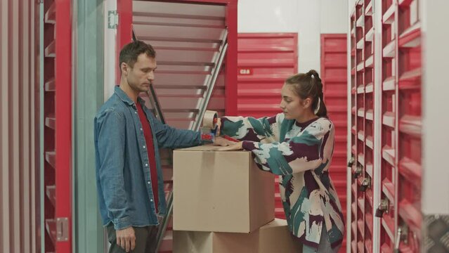 Young woman sealing box with adhesive tape while her husband putting it into locker room in self service storage