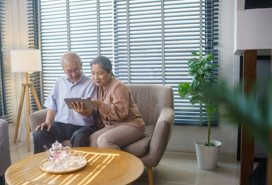 Asian grandfather and grandmother watching or meeting video call to family with digital tablet in the living room. Senior couple playing social media or searching on the internet.