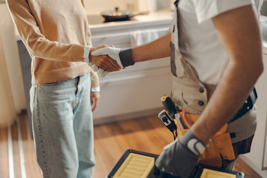 Close up of repairman in uniform shaking hands with woman while standing at home kitchen