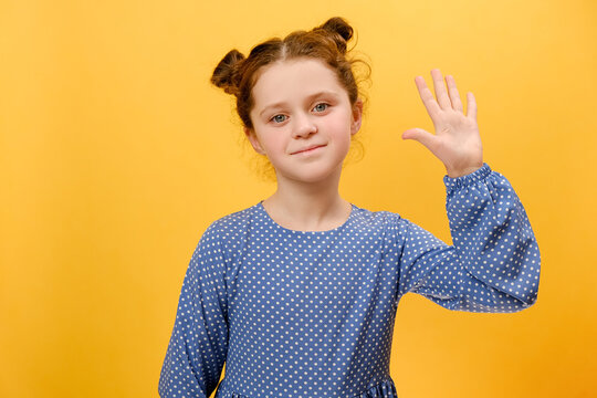 Portrait of preteen girl kid waving hand gesturing hello, smiling looking at camera, isolated on plain yellow color background wall in studio. Cute caucasian child showing hi gesture. Greeting concept