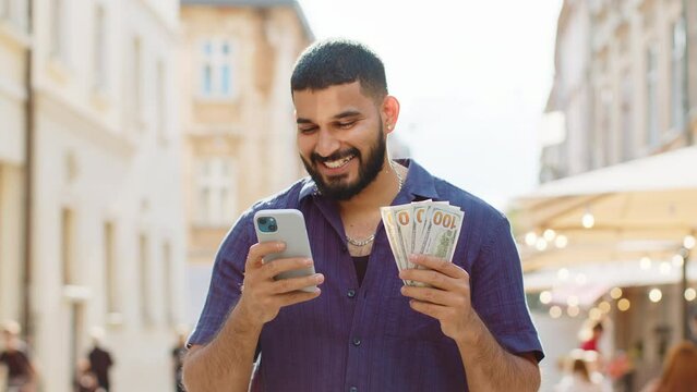Happy rich indian man counting holding money dollar cash, use smartphone calculator app, plans to order gifts and food delivery online, booking hotel room. Bearded guy tourist in city street, outdoors