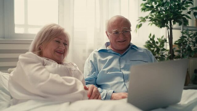 Happy senior couple lying in bed and watching a movie on laptop, weekend rest