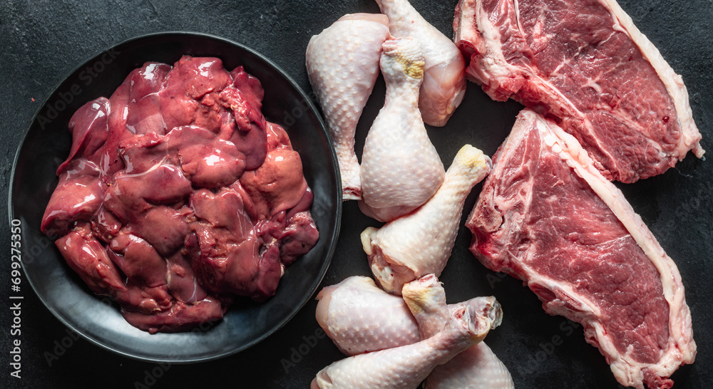 Wall mural Top view uncooked ingredients liver, chicken legs, and two beef steaks on a dark stone background