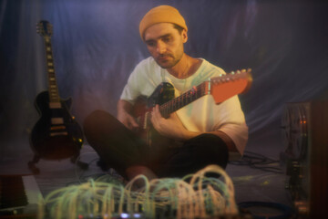 Blurry medium long shot of thoughtful adult male guitarist sitting legs crossed and playing electric guitar in makeshift studio