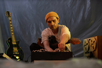 Medium shot of Caucasian male musician with stubble wearing yellow hat holding electric guitar switching cables on musical equipment in dim light studio