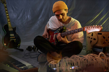 Medium long shot of male musician sitting legs crossed on studio floor and playing melancholic melody on electric guitar while recording with portable musical equipment