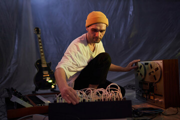 Medium shot of adult male musician sitting on makeshift studio floor and adjusting sound mixer board for recording onto magnetic tape recorder