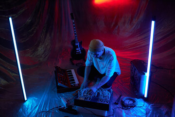 High angle shot of male composer working with sound mixer settings while squatting down makeshift studio with standing fluorescent lamps and plastic cover