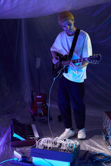 Vertical full length shot of adult male musician in blue neon light studio playing semi-acoustic guitar while using sound mixer for recording guitar samples
