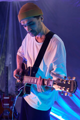 Vertical medium full shot of male band guitarist with stubble wearing beanie playing black semi-acoustic guitar on strap in blue neon light studio