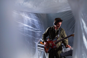 Medium long shot of male musician wearing jumpsuit and headphones adjusting portable musical equipment for electric guitar recording in studio covered with plastic