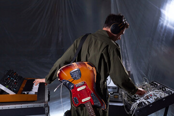 Back view of male sound producer with scuffed electric guitar behind back recording electric guitar samples with midi keyboard and audio mixer