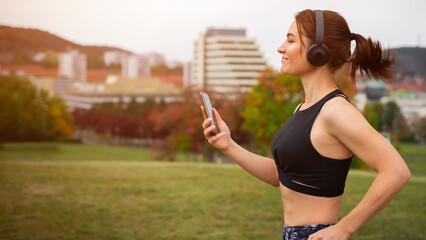 Wide photo of Athletic young woman with hair in a ponytail in leggings and a top with headphones is jogging on the background of the city. - 699286594
