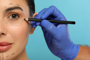 Doctor with pencil preparing patient for cosmetic surgery operation on light blue background, closeup