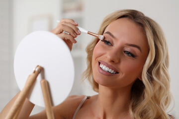 Beautiful makeup. Smiling woman applying eyeshadows in front of mirror indoors