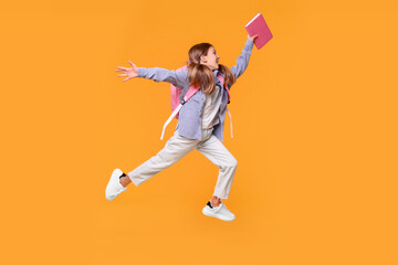 Fototapeta na wymiar Happy schoolgirl with book and backpack jumping on orange background