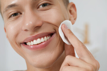 Handsome man cleaning face with cotton pad against blurred background, closeup