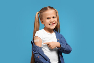 Happy girl pointing at sticking plaster after vaccination on her arm against light blue background