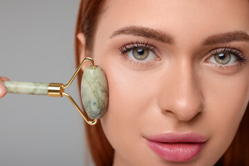 Young woman massaging her face with jade roller on grey background, closeup
