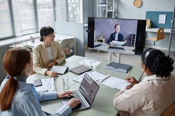 High angle shot of female business colleagues sitting at table in modern office and looking at plasma screen while having online video call meeting - obrazy, fototapety, plakaty