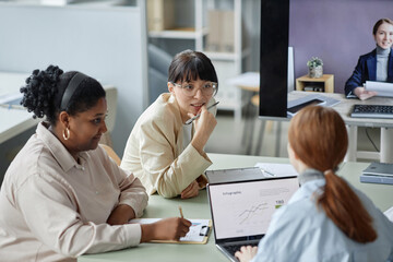 Medium shot of company employees sitting at table in office during video conference call, focus on brunette business lady propping up chin while discussing work with diverse colleagues - obrazy, fototapety, plakaty