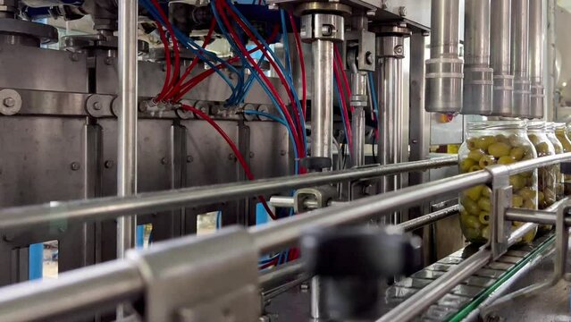 Close-up of the movement of jars filled with olives on the conveyor belt. Glass jars of green olives move on an automatic conveyor belt in a cannery. Automatic marinade pouring. Food industry.