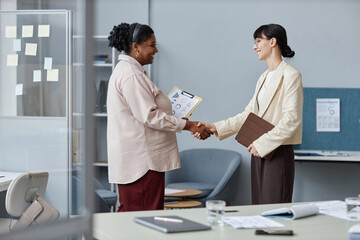 Medium full shot of two smiling diverse business women shaking hands while standing in modern office