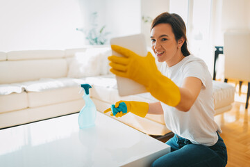 The young woman is demonstrating on her tablet how she cleans the living room wearing yellow gloves