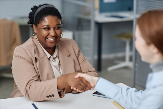 Medium shot of cheerful African American female HR agent shaking hands at desk with unrecognizable job candidate in company office