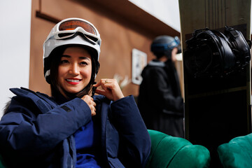 Female asian traveler sits on hotel lobby couch, wearing winter jacket and adjusting her ski...