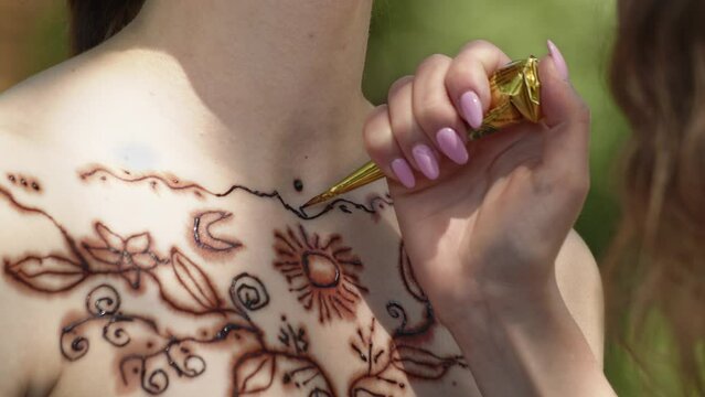 A young girl in black dress makes henna tattoo on chest with an Indian oriental pattern. A woman professional tattoo artist applies a drawing with a special tool. Close up. 