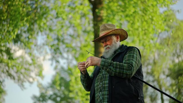 Old man wearing sunhat and preparing supplies for fishing at forest. Grandfather angler putting hook bait to his fish rod. Pensioner spending his weekend at the lake pier. Concept of relaxation