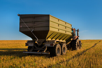 A grain trailer is attached to a tractor, in a field with stubble and a blue sky