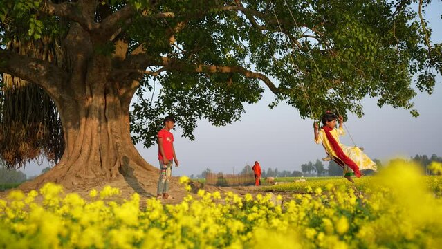 Swinging beneath a majestic banyan tree, children laugh and play amidst a vast golden mustard field. Their carefree joy paints a vibrant picture against the serene rural backdrop