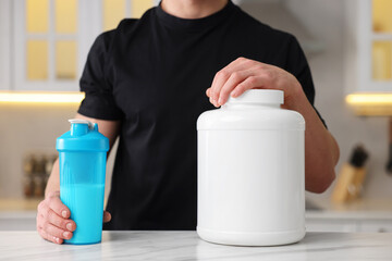 Young man with shaker of protein and powder at white marble table in kitchen, closeup